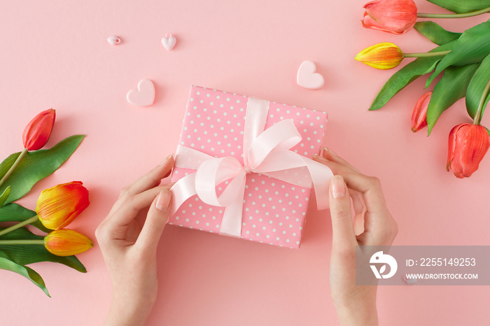 Spring gift concept. First person top view photo of female hands holding gift box with silk ribbon bow and tulips flowers on pastel pink background. Spring holiday idea.