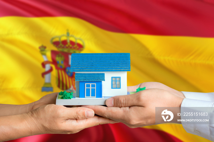 Spain real estate concept. Man and woman holding miniature house in hands. Citizenship theme and national flag on background.