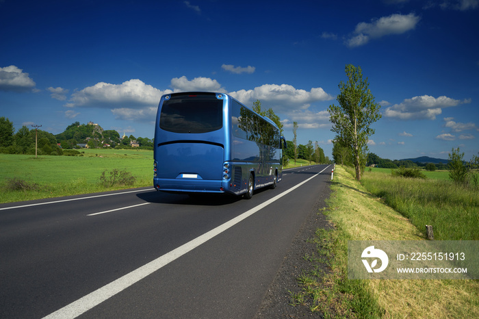 Blue bus traveling on asphalt road in a rural landscape. Village and mountain with castle ruins in the background.