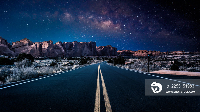 An endless desolate road leading into Arches National Park in Moab, Utah, USA under a dark and starry night sky.