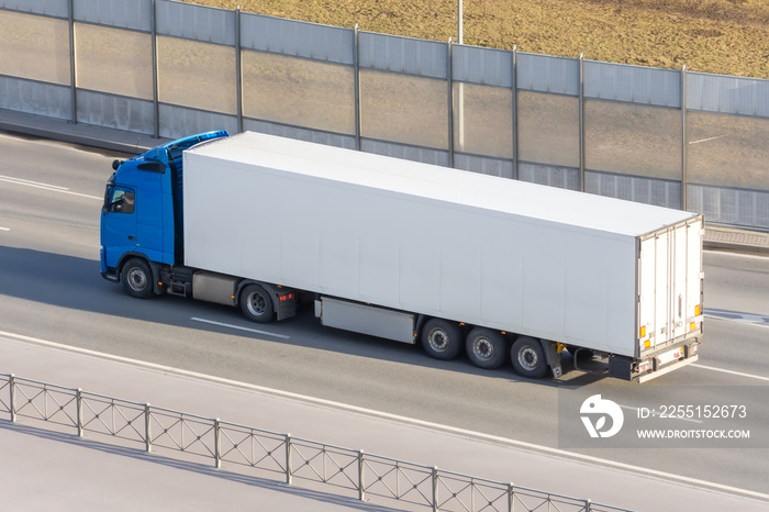 White truck with gray awning trailer rides on a city highway, side aerial view.