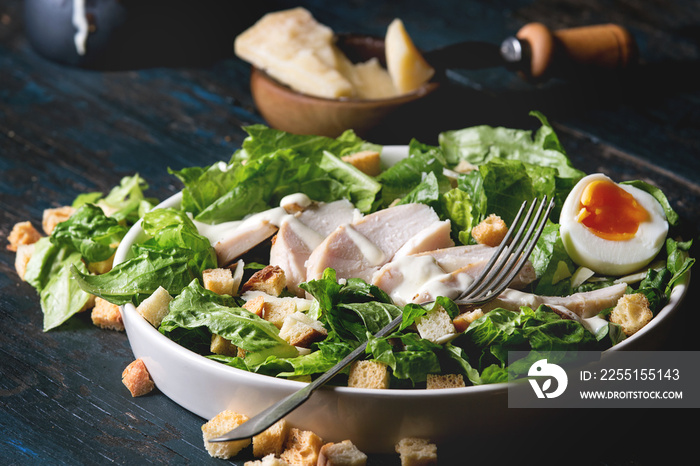 Classic Caesar salad with grilled chicken breast and half of egg in white ceramic plate. Served with fork and ingredients above over old dark blue wooden background. Rustic style