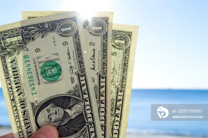 Person holding in hand paper bills of dollars with face value of one dollar on background of brightly shining sun, blue sky and calm blue sea. One-dollar paper banknotes on backdrop of blue sea sky