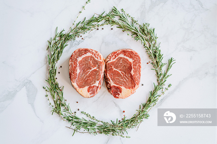 raw two ribeye steaks in the center of a Christmas wreath of rosemary thyme cherry tomatoes and bell peppers on a white marble surface. christmas dinner concept for two