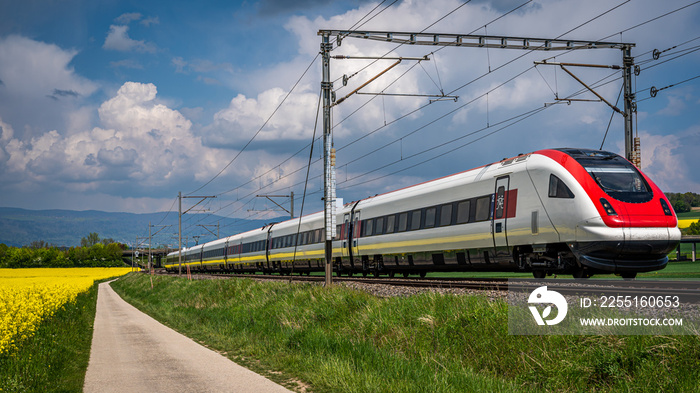 Train running through rapeseed field in spring. Red train, railroad with blue sky, yellow flowers and green field. Chavornay, Switzerland.