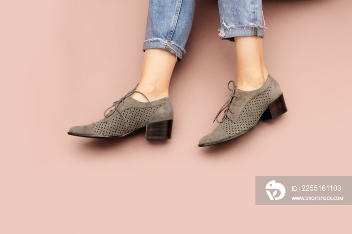 Woman’s Caucasian legs in blue twisted jeans and gray suede English brogue Oxfords shoes isolated on light pink beige background. Casual sneakers. Front top view, flatlay, copy space