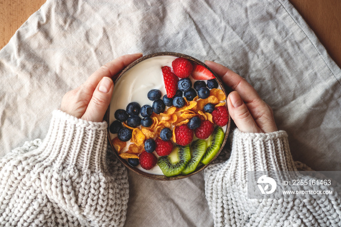 Healthy breakfast in coconut bowl on table with linen tablecloth. Yogurt with corn flakes and blueberry, strawberry, kiwi and raspberry. Womans hand wearing sweater. Vegetarian food