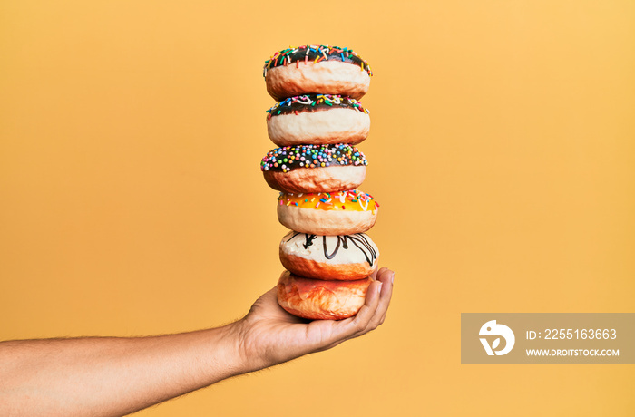 Hand of hispanic man holding tower of donuts over isolated yellow background.