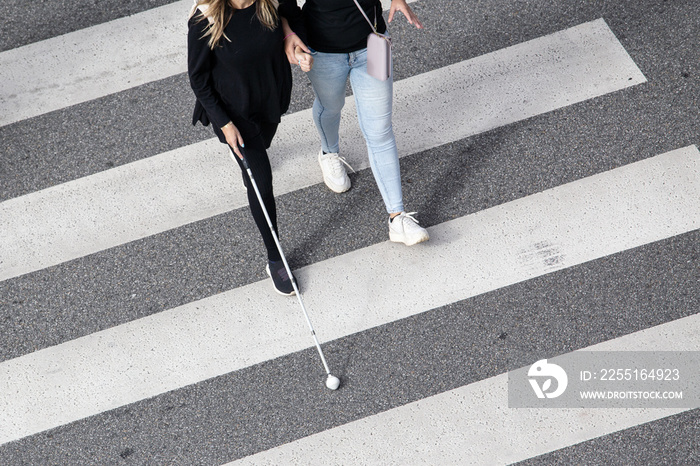 Scene of a Blind woman walking on zebra crossing helped by another person using her white cane. Help in the early stages of blindness