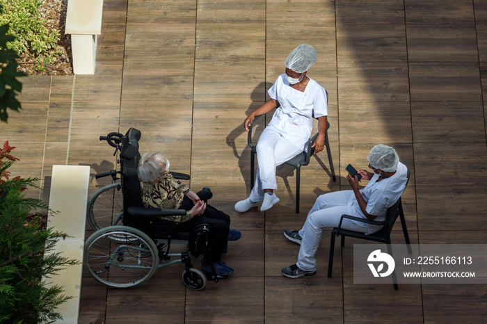 Nurses caring for elderly or disabled people in a nursing home during the coronavirus pandemic Covid-19. Carers having a break in th sun with an old woman resident.