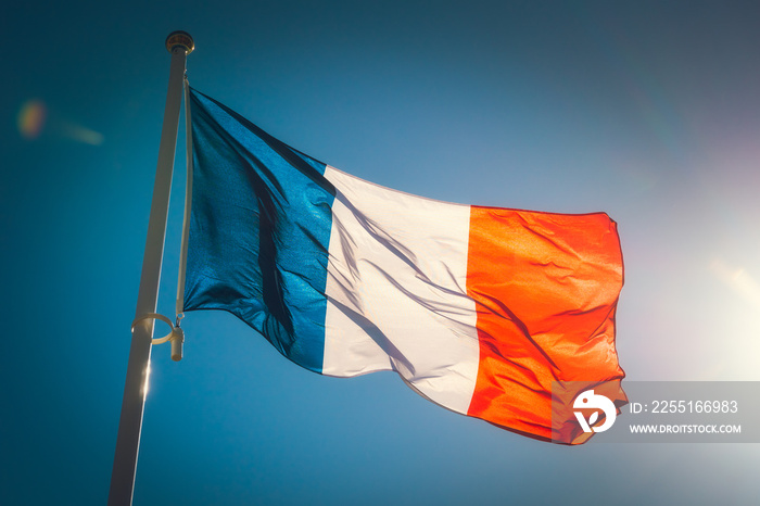 Looking up to a beautiful waving french flag against a blue sky with sunlight.