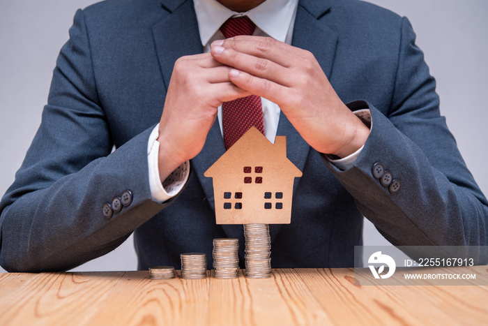 Businessman protecting wooden house model with growing stack of coins, business financial and investment concept