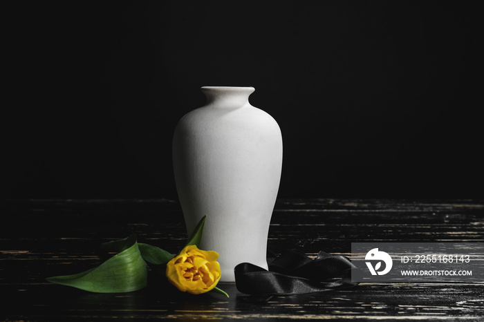 Mortuary urn with ribbon and flower on table against dark background