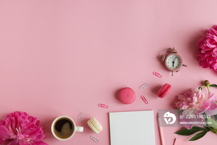 Business and education concept. Feminine workplace with notepad, pencil, alarm clock, peony flowers, a cup of coffee and macarons on a pink background with copy space. Top view, flat lay