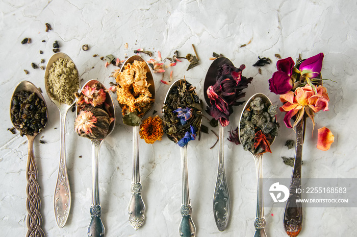 Assortment of dry green herbal and flower tea in a spoons on a white stone background.