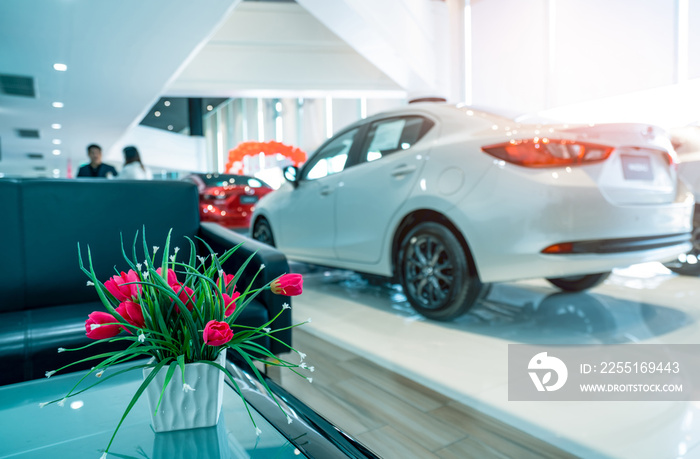 Fake red flowers in ceramic white vase on blurred background of sale person deals with customer in modern showroom. Car dealership. Blurred luxury car parked in showroom. Automotive business industry.