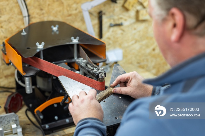 Industrial tool worker grinds a steel plate on a rotating belt sander, he makes a knife.