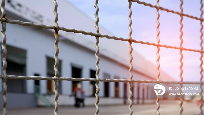 Focus on steel mesh fence in foreground with flare light and blurred background of large distribution warehouse, security in business industrial concept
