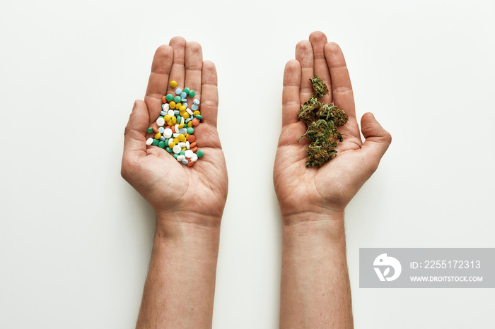 Top view of colorful pills in one hand and medical marijuana buds in another over white background. Concept of choice and herbal, alternative medicine