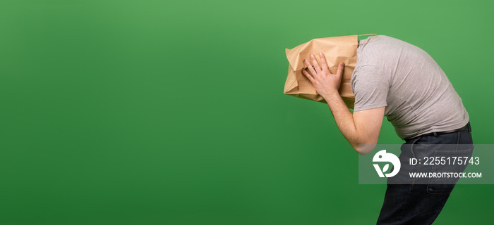 An unrecognizable man with a paper bag on his head grabbed his head against a green background. Place for your text. mental health, greenwashing, disinformation concept