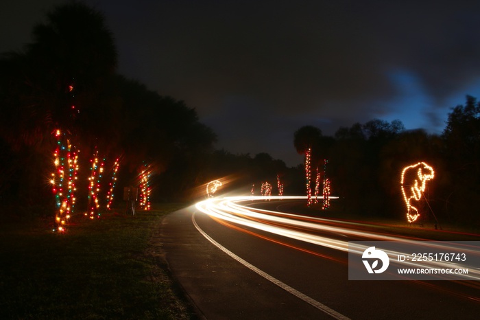 Cars pass by a colorful display of illuminated Christmas decorations, their lights creating streaks at twilight in a slow shutter speed time exposure