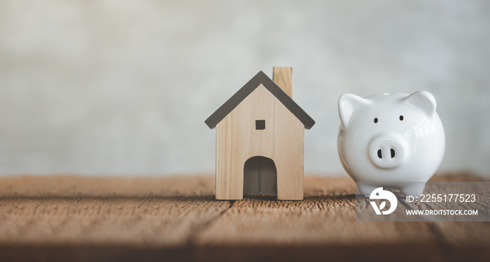 White piggy bank with wooden house on wood table and blurred copy space background. Concept for financial home loan or money saving for house buying.