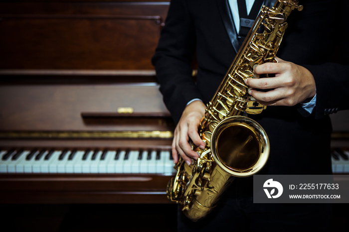 close up of Young Saxophone Player hands holding alto sax musical instrument with piano background in dark room, vintage tone, can be used for music background, copy space