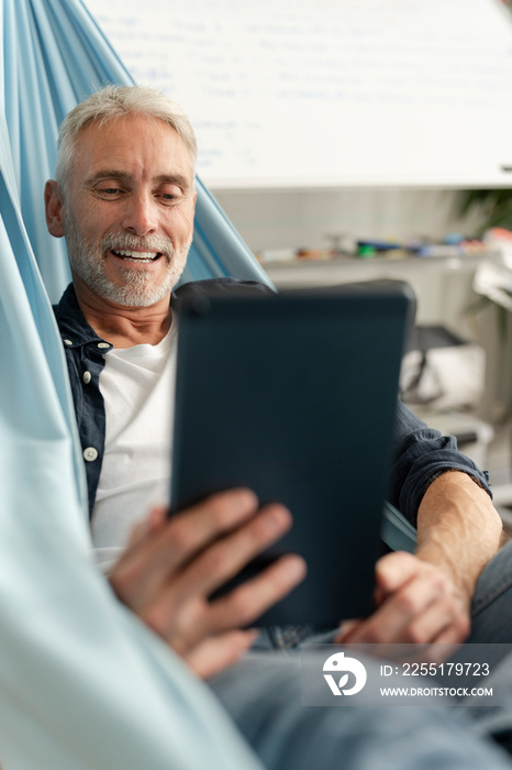 Smiling mature man lying in hammock and using digital tablet