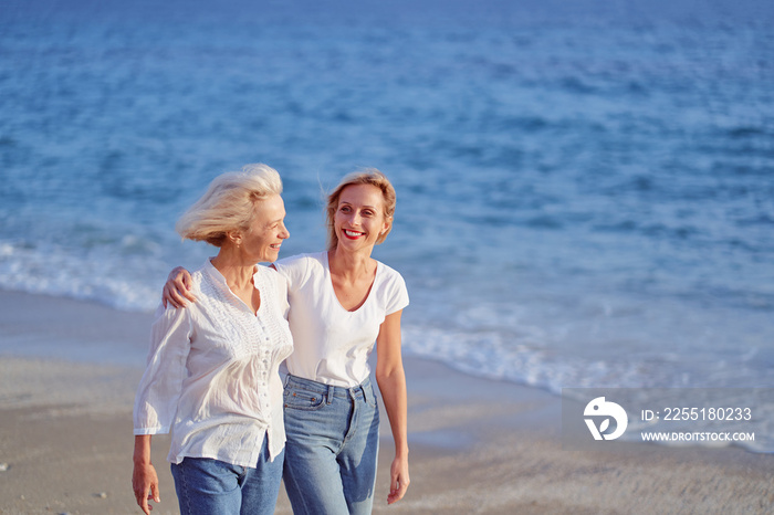 Outdoor portrait of smiling happy caucasian senior mother with her adult daughter hugging and walking on sea beach.