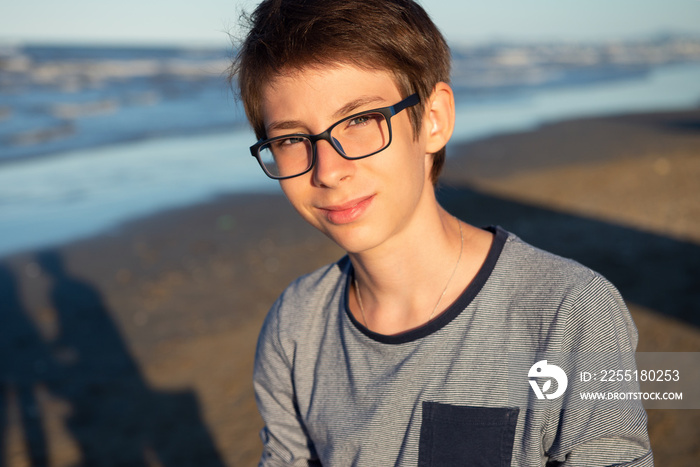 Young boy posing at the summer beach. Cute spectacled smiling happy 12 years old boy at seaside, looking at camera. Kid’s outdoor portrait over seaside.