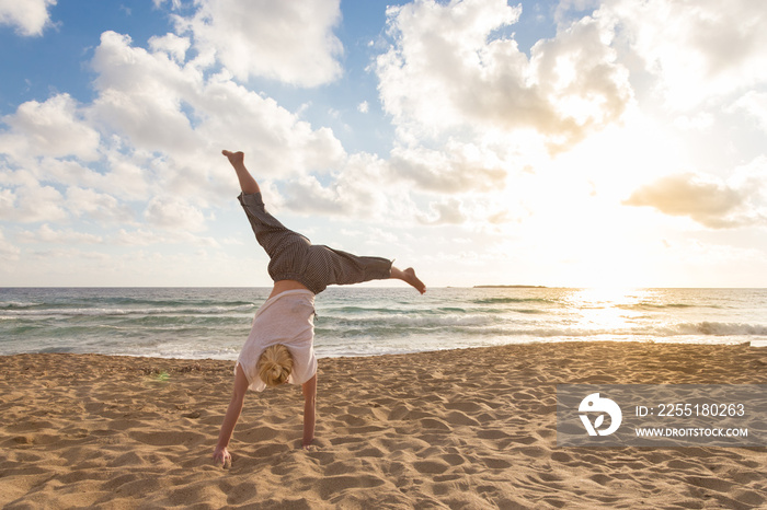 Relaxed woman enjoying sun, freedom and life turning cartwheel on beautiful beach in sunset. Young lady feeling free, relaxed and happy. Vacations, freedom, happiness, enjoyment and well being.