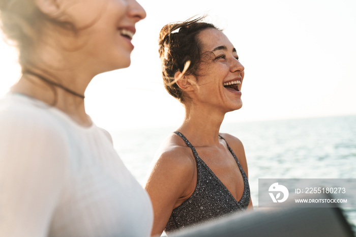 Image of happy fitness women smiling and standing with yoga mat by seaside in morning