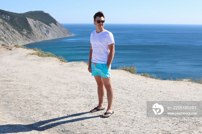 Smiling looking sideways young caucasian man with sunglasses in white t-shirt and blue shorts on mountain on sea blurred sunny background. Portrait in full growth of a 32 year old young man.