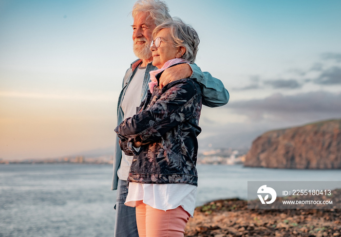 Lovely senior couple standing on the rocks at sea enjoying nature and freedom looking the horizon at sunset light. Relaxed lifestyle for a caucasian couple of retirees