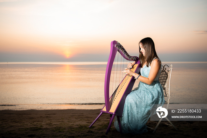 A girl in a flower dress plays on a Celtic harp by the sea at sunset