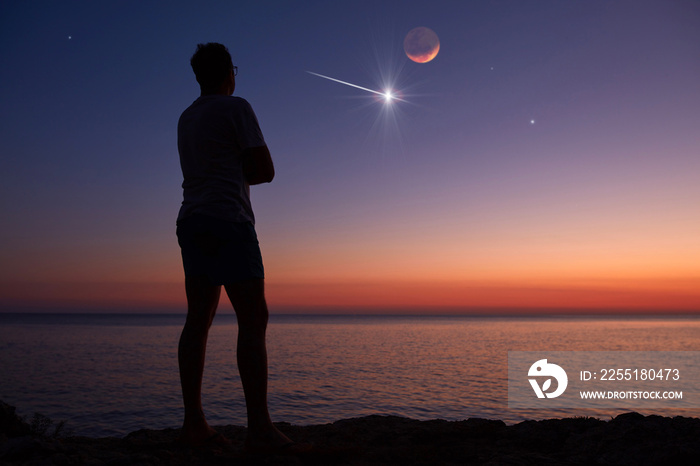 Silhouette of a man looking at the Moon and stars over sea ocean horizon.