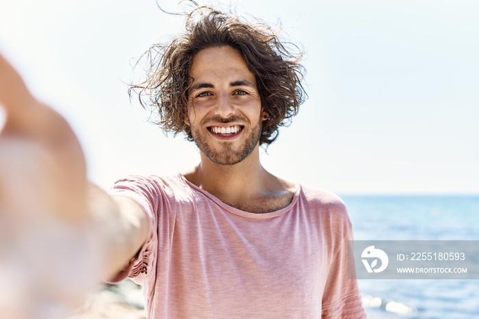 Young hispanic man smiling happy make selfie by the camera standing at the beach.