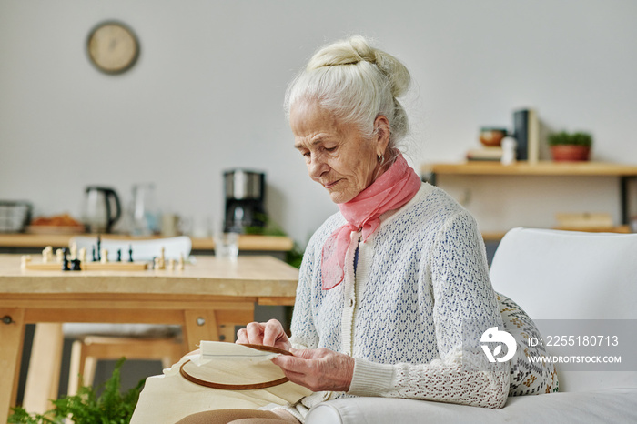 Elderly woman concentrating on her handicraft, she sitting on armchair and embroidering