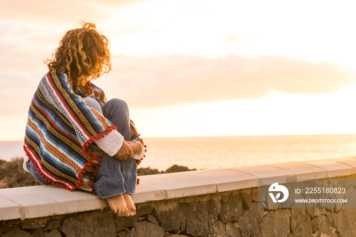 Unrecognizable lonely woman sit down on a wall looking and enjoying the sunset on the sea - coloured trendy mexican poncho style and people in outdoor leisure relax activity