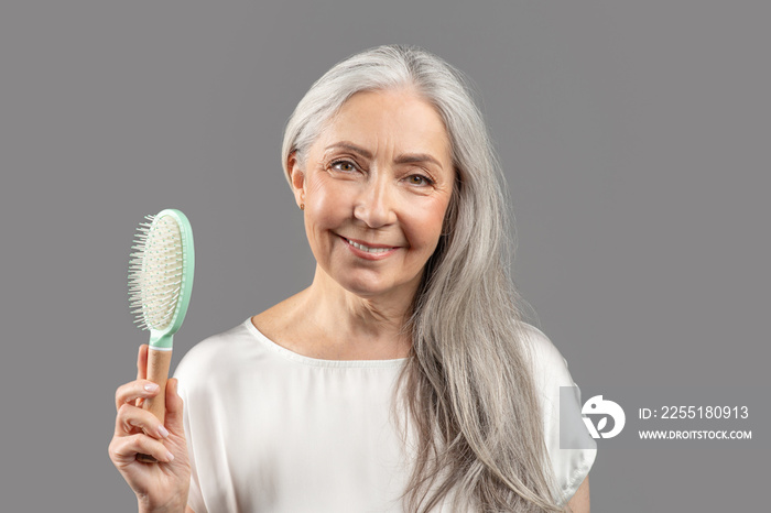 Aging gracefully. Charming senior lady with silky long hair holding brush and smiling on grey studio background