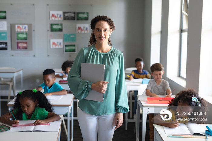 Portrait of caucasian young female teacher standing by multiracial students studying at desk