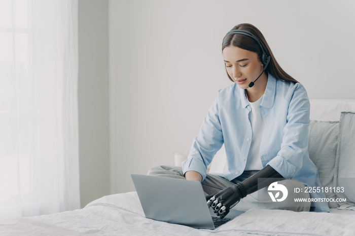 Disabled girl in headset typing on laptop using bionic prosthetic arm, sitting on bed at home