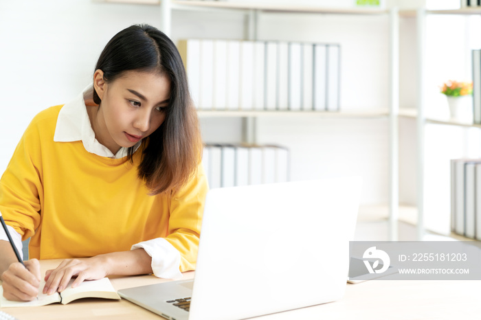 Young attractive asian female student sitting at table looking at laptop writing journal by hand note idea script, diary or sketch design on notebook at home office or library with copyspace concept.