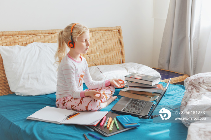 Caucasian girl child meditating in bed and learning online on laptop Internet. Virtual class lesson on video during home self isolation. Distant remote video education. Modern school study for kids