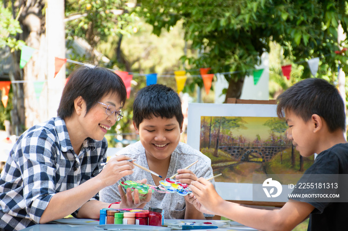 Asian middle-aged female art teacher is encouraging and explaining the process of watercolor painting to young asian boys during their art lesson in the school park, raising teens concept.