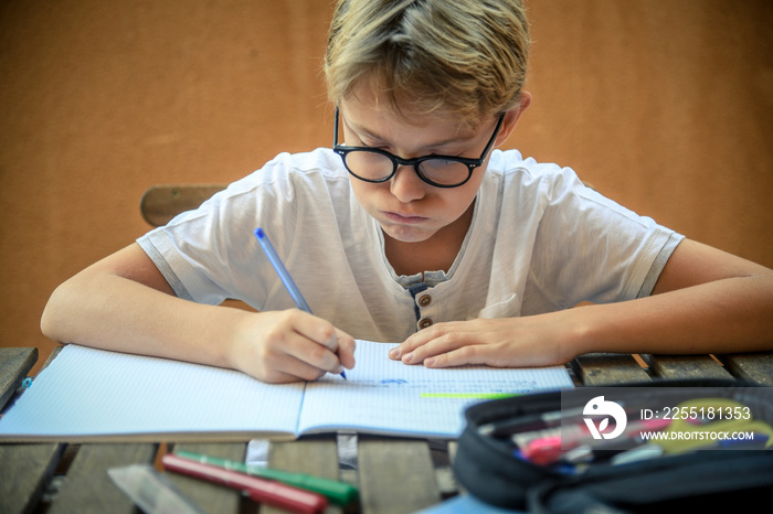 Cute blond child with glasses sitting at the table doing homework for school Young student intent on studying Boy concentrated in the study Fatigued and bored, snort Writing on an exercise book