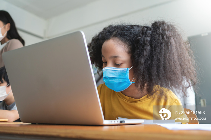 Adorable Black African American girl wearing protective face mask using laptop for studying in classroom at international school.