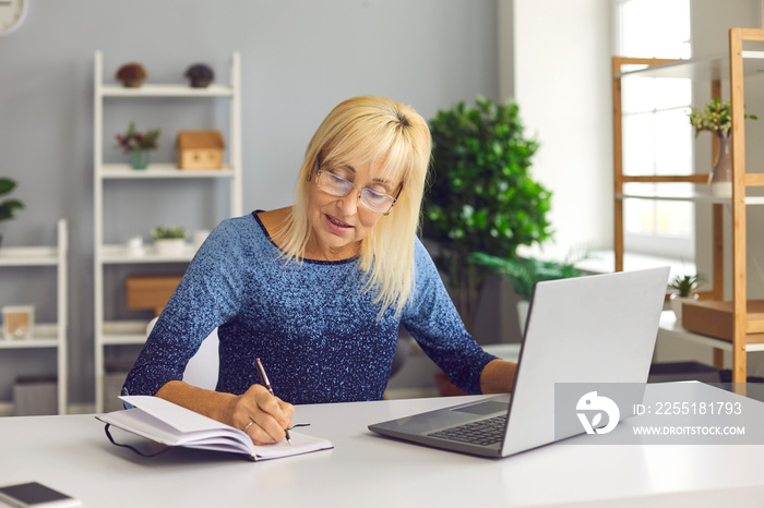 Senior woman making notes in notebook while looking at business training or online webinar on laptop. Concept of remote work or social distance learning at home.