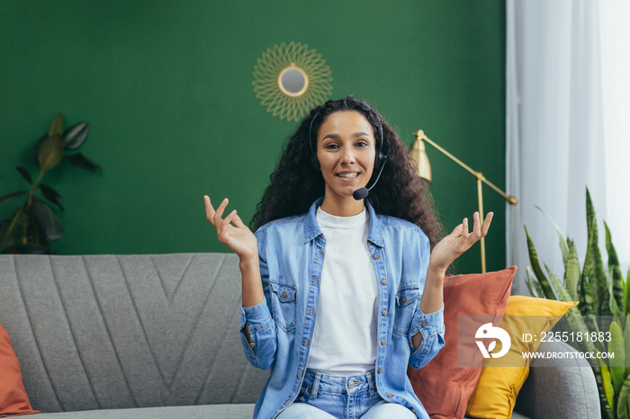 Joyful hispanic woman with video call headset at home smiling and looking at camera, woman studying and working online remotely sitting on couch at home.