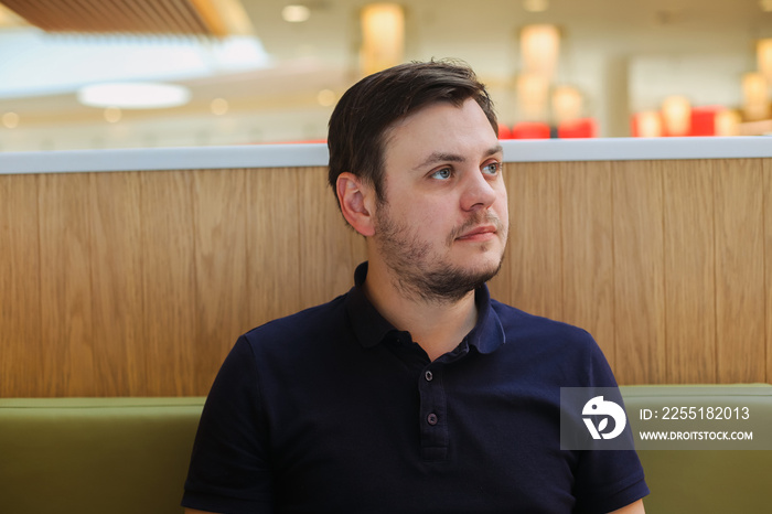 looking away young 32 years old caucasian unshaven beard man in a dark blue polo shirt in cafe at the food court. Waiting concept, portrait